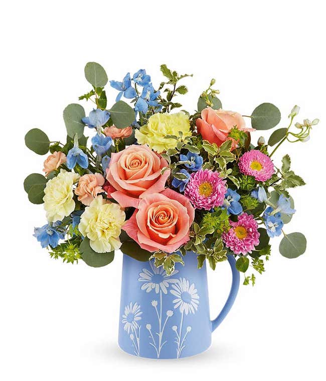 Floral arrangement of Peach Roses, Yellow carnations, peach mini carnations, light blue delphinium, and floral greenery in a pale blue pitcher vase with white daisy details