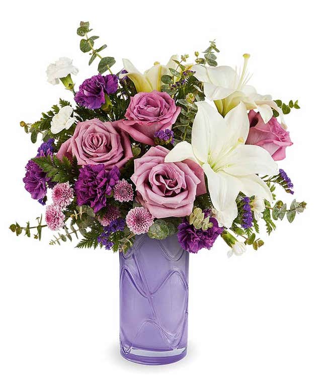 Lavender Roses, White Asiatic Lilies, Purple Carnations, Mini White Carnations, Lavender Button Poms, and Floral Greenery in a tall purple glass vase with a wave design