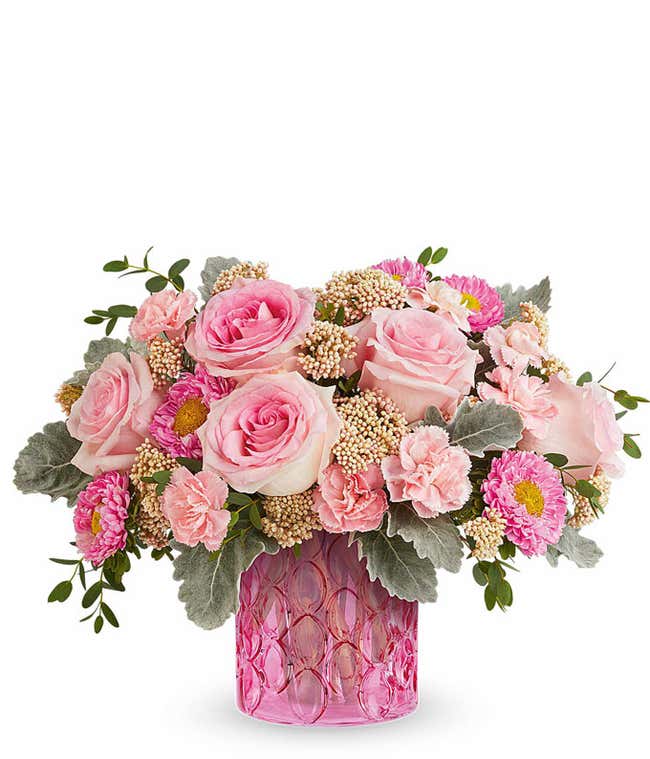 Pink floral arrangement with roses, spray roses, carnations, and Matsumoto asters in a textured pink glass vase with greenery.