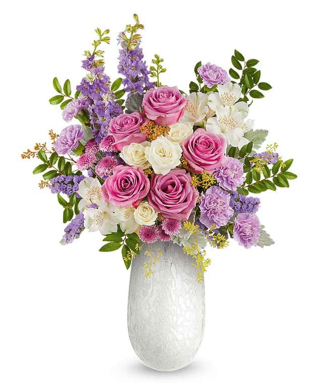 Lavender roses, white spray roses, white alstroemeria, lavender carnations, lavender larkspur, and lavender button poms with floral greenery in a white pearlescent vase