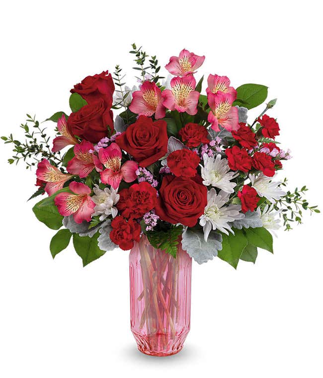 Valentine&rsquo;s Day floral arrangement featuring red roses, red carnations, pink alstroemeria, white cushion mums, and lush greenery arranged in a pink glass vase.