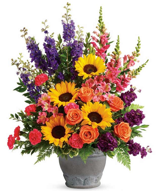 A sympathy arrangement featuring Orange Roses, Pink Alstroemeria, Hot Pink Carnations, Yellow Sunflowers, Purple Larkspur, Hot Pink Snapdragons, Purple Stock, and Floral Greenery in a Grey Ceramic Pot, offering comfort and solace during times of sorrow.