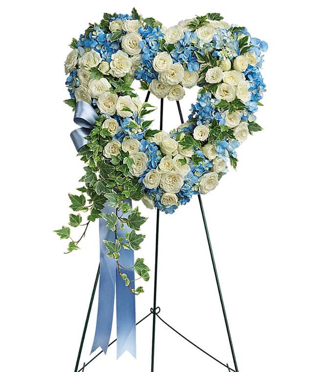 Heart-shaped funeral spray with white roses, blue hydrangeas, green ivy, and a blue ribbon on a metal easel.