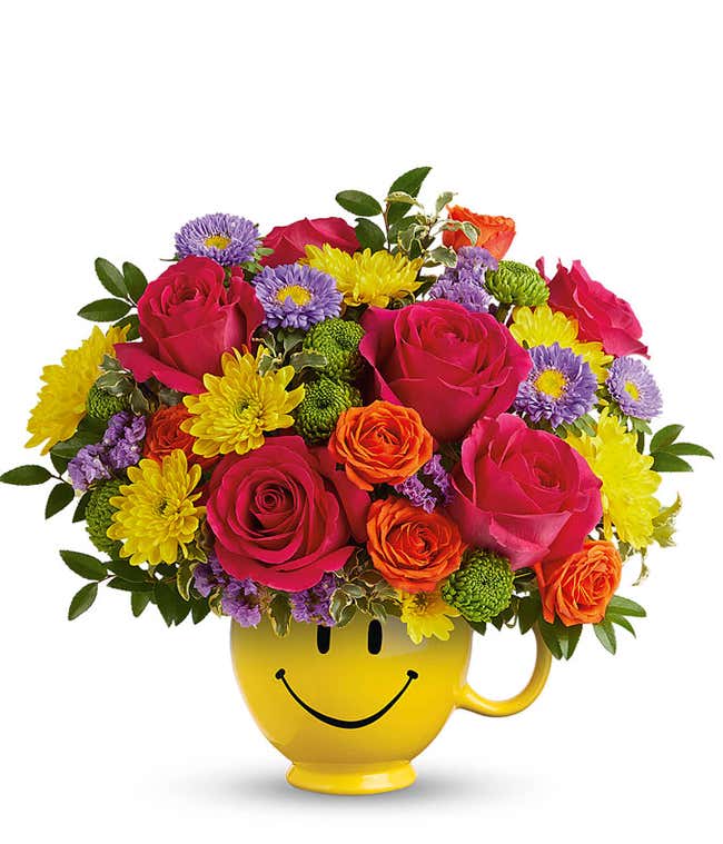 Hot Pink Roses, Orange roses, lavender asters, Yellow &amp; green chrysanthemums, floral greens in a yellow smiley face mug vase against a white background
