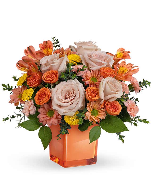 Light orange and coral roses, peach alstroemeria, peach carnations, Yellow and bronze chrysanthemums, pravifolia eucalyptus, lemon leaf in a glass coral cube vase against a white background