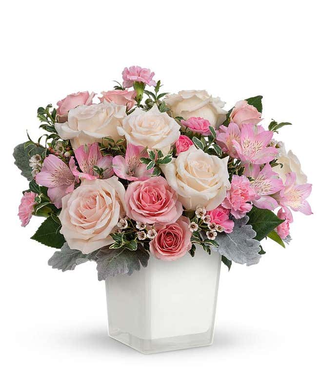 A floral arrangement featuring cr&egrave;me roses, pink spray roses, pink alstroemeria, pink miniature carnations, white waxflower, and greenery in a white vase.