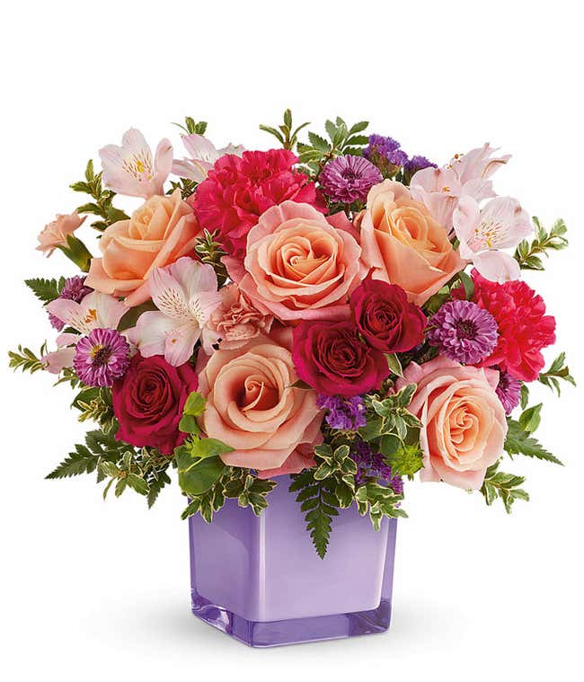Peach Roses, hot pink spray roses, light pink alstroemeria, hot pink carnations, mini peach carnations, purple button poms, and floral greenery in a purple cube vase