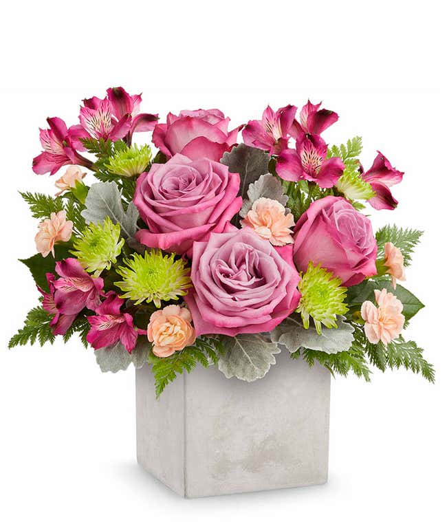 Floral arrangement with lavender roses, purple alstroemeria, mini peach carnations, green button poms, and floral greenery in a grey faux concrete look cube