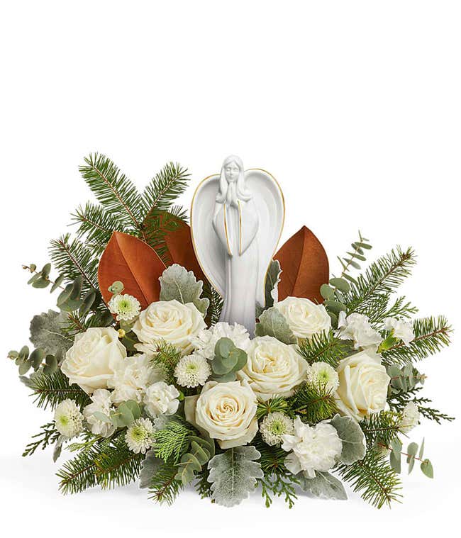 Floral centerpiece featuring white roses, white chrysanthemums, silver-toned foliage, magnolia leaves, and evergreen sprigs, with a peaceful angel figurine at the center.