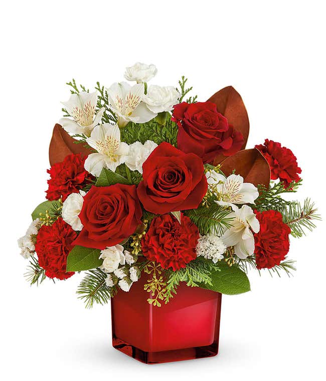 Holiday floral arrangement featuring red roses, red carnations, white alstroemeria, and white miniature carnations, accented with greenery, brown magnolia leaves, and evergreen sprigs, arranged in a red cube vase.