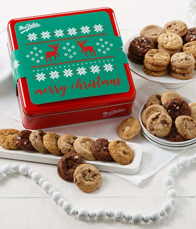 Red holiday cookie tin with a green lid featuring white snowflakes, red reindeer, and the words &ldquo;Merry Christmas,&rdquo; surrounded by plates of assorted bite-sized cookies including chocolate chip, triple chocolate, and white chocolate macadamia.