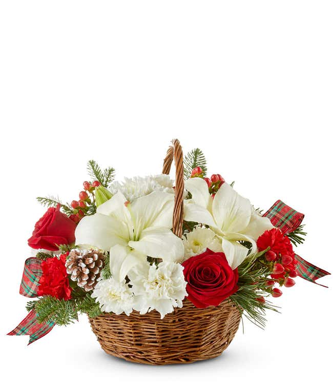 A brown woven basket filled with white lilies, red carnations, red roses, white poms, red berries, and christmas greens with a handle. 