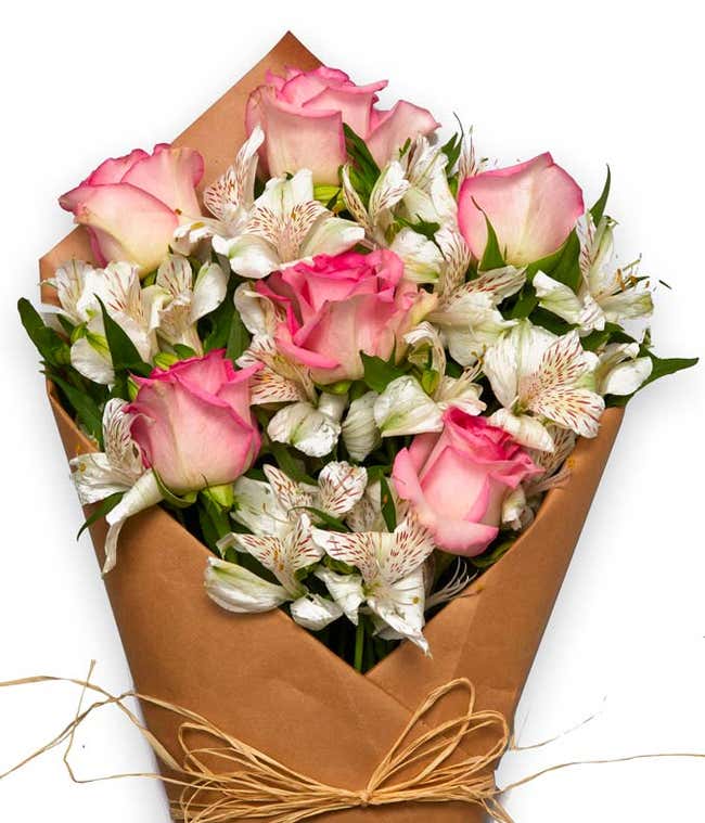 A bouquet featuring pink roses, white alstroemeria, and fresh greens, wrapped in brown parchment paper and tied with twine.