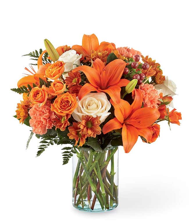 Clear glass vase filled with orange lilies, white roses, orange spray roses, peach carnations, orange daisy poms, red hypericum berries, and greenery.