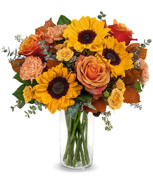 Fall bouquet with sunflowers, orange roses, peach carnations, yellow spray roses, seeded eucalyptus, and autumn oak leaves in a tall clear glass vase.