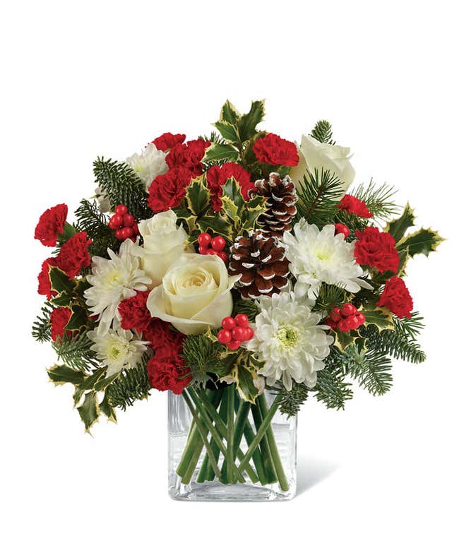 Holiday floral arrangement with white roses, red miniature carnations, white chrysanthemums, pinecones, red berries, holly leaves, evergreen sprigs, and a clear glass cube vase.
