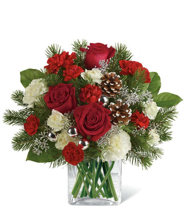 A festive arrangement with red roses (deluxe &amp; premium only), red carnations, white carnations, red mini-carnations, white mini-carnations, seasonal greens, silver ornaments, and pine cones.