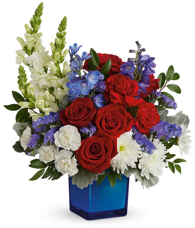 Memorial Day arrangement featuring red roses, red carnations, white miniature carnations, blue delphinium, white snapdragons, white cushion chrysanthemums, and lush floral greenery in a blue glass cube vase.