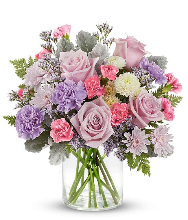 Soft pastel floral arrangement featuring lavender roses, lavender carnations, pink miniature carnations, lavender cushion spray chrysanthemums, white button spray chrysanthemums, dusty miller, and lush greenery in a short cylinder vase.