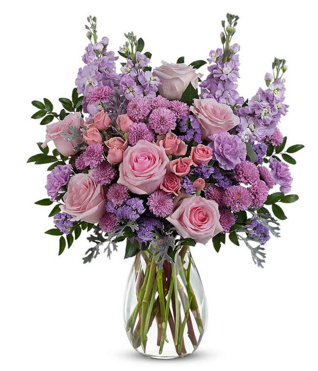 Pink roses, pink spray roses, lavender carnations, lavender stock, lavender button spray chrysanthemums, lavender sinuata statice, and floral greenery arranged in a clear glass jordan vase.