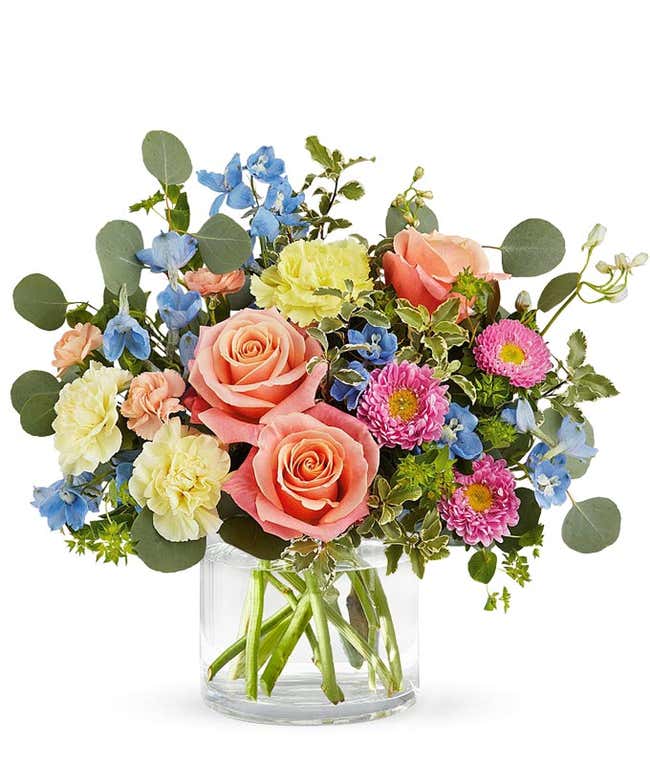 A bouquet of peach roses, yellow carnations, peach mini carnations, blue delphinium, and pink matsumoto asters with greenery in a short clear cylinder vase