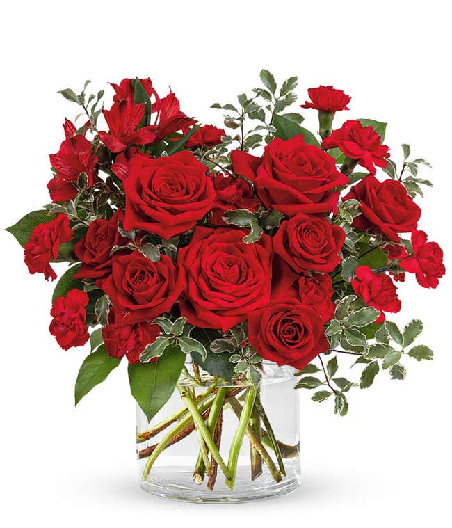An all red bouquet with the blooms arranged in the shape of a heart consisting of roses, spray roses, alstroemeria, mini carnations, and greens in a short cylidner vase