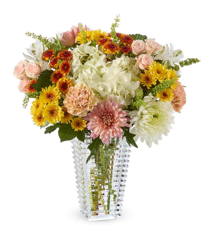 A bouquet in a textured rectangular vase of bronze, gold, peach and white mums, white alstroemeria, peach carnations, pink spray roses, white hydrangeas and greens.