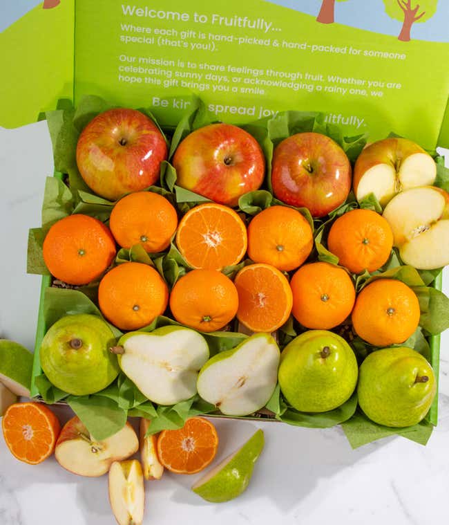 A green box filled with apples, pears, and mandarins, all arranged on green tissue paper with some fruits cut open to display their fresh interior.