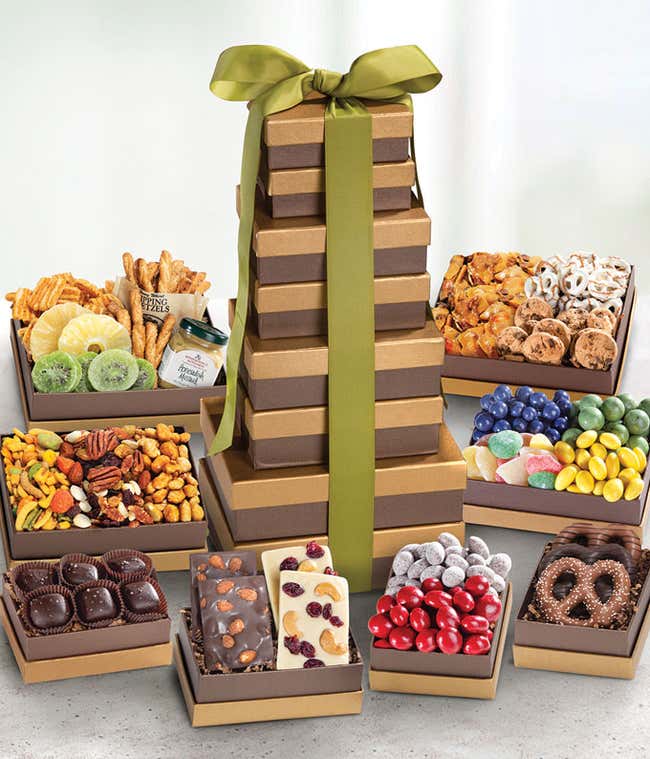 A tall 8-box gift tower of brown and gold boxes tied with a green ribbon. Surrounding the tower are trays filled with dried kiwi, nuts, chocolates, pretzels, and more, arranged for a vibrant, abundant display.