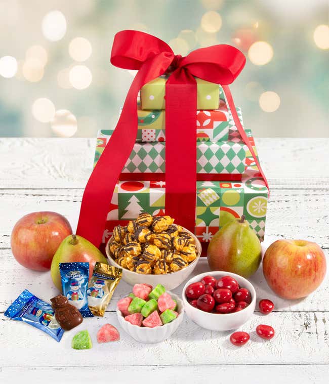 A festive stack of gift boxes wrapped with a red bow, filled with apples, pears, caramel popcorn, chocolate cherries, and red and green gummy candies. 