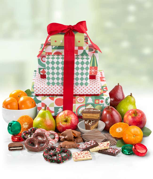 A multi-tiered holiday tower decorated with red ribbon.  It includes fresh oranges, pears, and apples, alongside chocolate-dipped pretzels, peppermint bark, and other holiday-themed chocolates and sweets. 