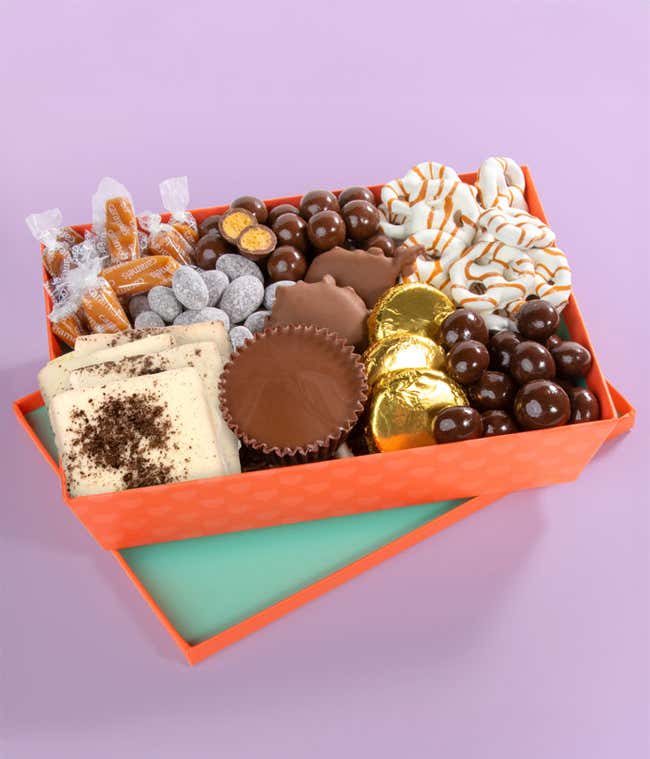 Gift box filled with chocolate-covered almonds, chocolate-covered pecan patties, chocolate cups, and chocolate sandwich cookies wrapped in gold foil, white chocolate-covered pretzels, milk chocolate-covered candy, and a few caramel candies.