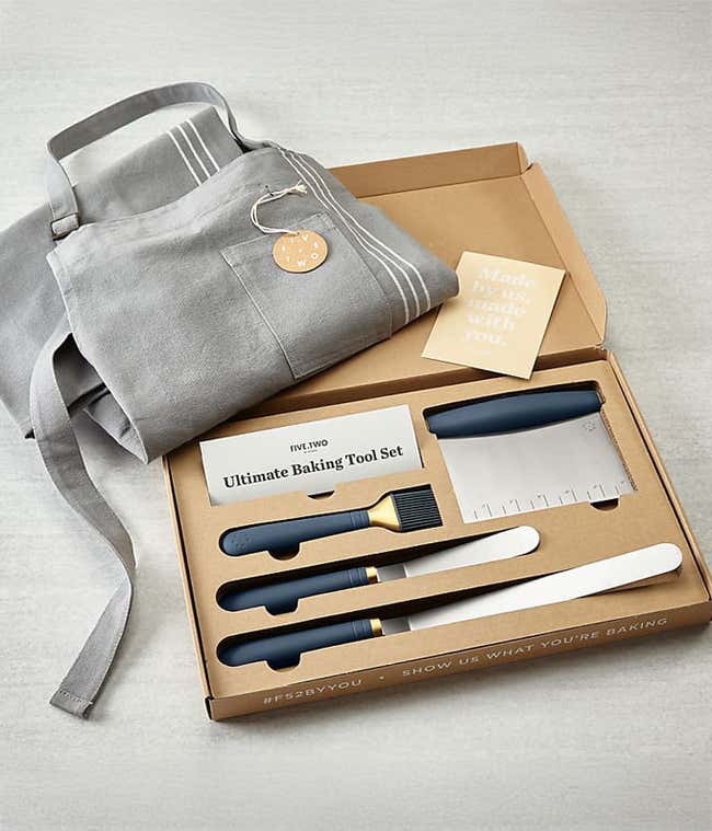 Gray linen apron and baking tool set displayed in a cardboard box. The set includes a dough scraper, a pastry brush, a spatula, a bench scraper, and an offset spatula. The apron is neatly folded and features a simple striped design.