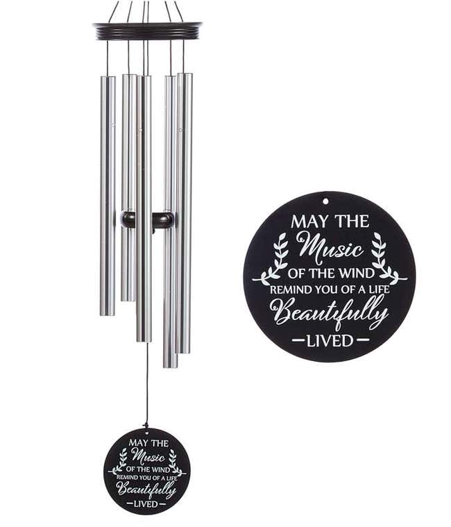 A windchime with metal rods and a wooden accents and sail. Black sail with white lettering reads may the music of the wind remind you of a life beautifully lived.