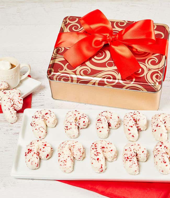  A festive red and gold tin with a large red bow, filled with candy cane-shaped cookies coated in white icing and sprinkled with crushed peppermint. The tin is displayed next to a tray of the same cookies, with a small cup of hot cocoa topped with marshma