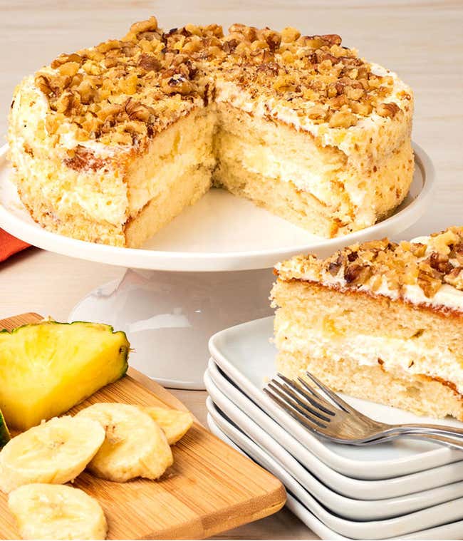 A layered hummingbird cake topped with chopped walnuts and light frosting on a white cake stand. A slice on stacked plates shows the moist interior with pineapple and banana. Fresh pineapple and banana slices are on a wooden board beside the cake.