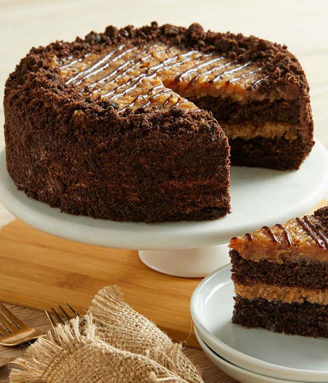  A rich German chocolate cake on a white cake stand, featuring layers of moist chocolate cake filled with a coconut-pecan frosting. The cake is topped with a generous layer of the same frosting and a drizzle of chocolate. A slice is served on a plate besi