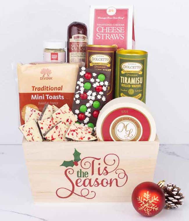 A wooden basket with the words Tis the Season, filled with sausage, cheese, brie, mustard, cookies, toasts, rolled wafers, and festive peppermint bark, accented by a holiday ornament and pinecone.