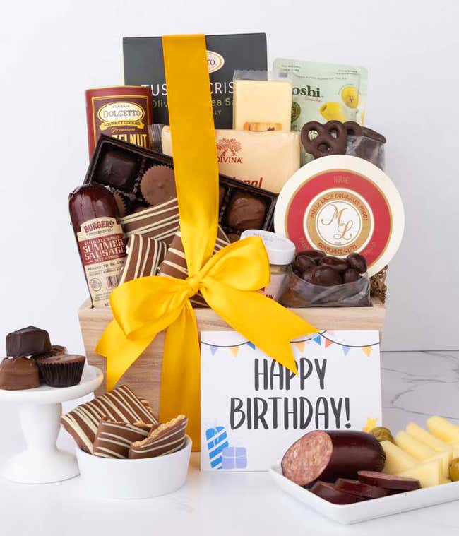 A wooden basket filled with chocolates, summer sausage, cheeses, crackers, crisps, mustard, and snacks, with displays of chocolate-covered treats, sliced sausage, and cheese styled for a Happy Birthday message card.