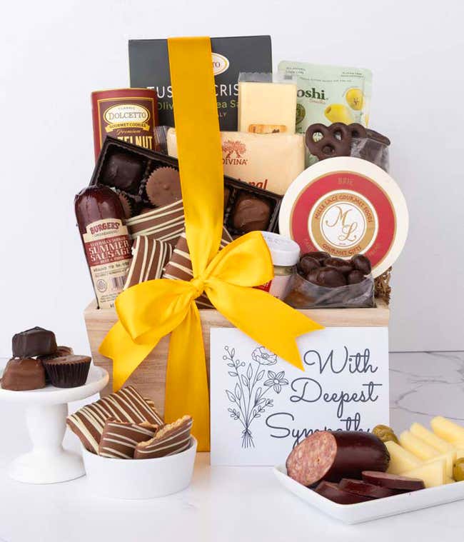 A wooden basket filled with chocolates, cheeses, sausage, crackers, crisps, and snacks, displayed with a With Sympathy card, chocolate treats, and a plate of sliced sausage, cheese, and olives.