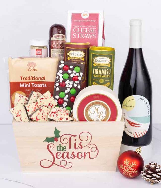 A wooden basket with the words Tis the Season, filled with sausage, cheese, mustard, cookies, wafers, and holiday bark, paired with a bottle of Hill &amp; Harbor Pinot Noir beside a red ornament and pinecone.
