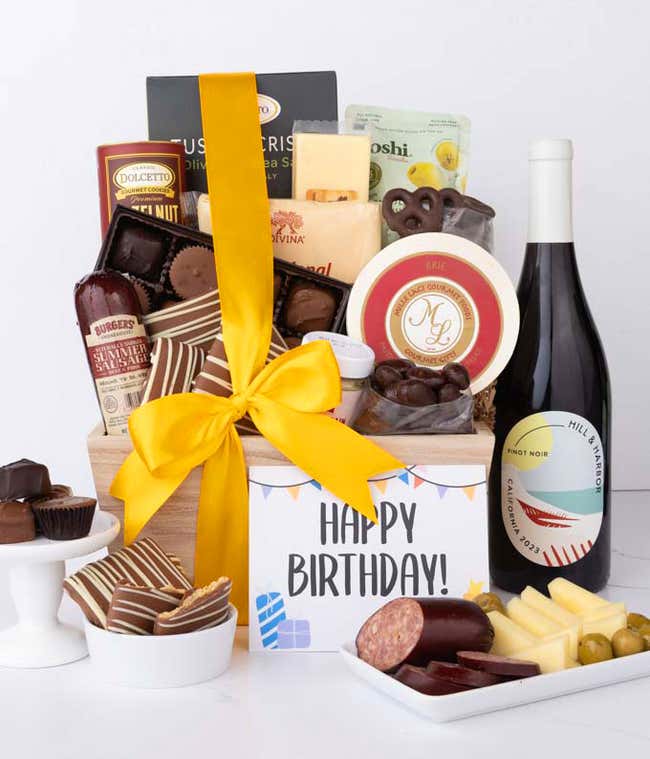A wooden basket filled with chocolates, summer sausage, cheeses, crackers, crisps, mustard, and snacks, with displayed next to a bottle of wine chocolate-covered treats, sliced sausage, cheese and a Happy Birthday message card.