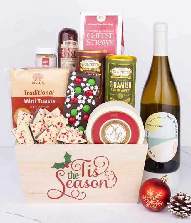 A wooden basket with the words Tis the Season, filled with sausage, cheese, mustard, cookies, wafers, and festive bark, paired with a bottle of Hill &amp; Harbor Chardonnay, next to a red ornament and pinecone.