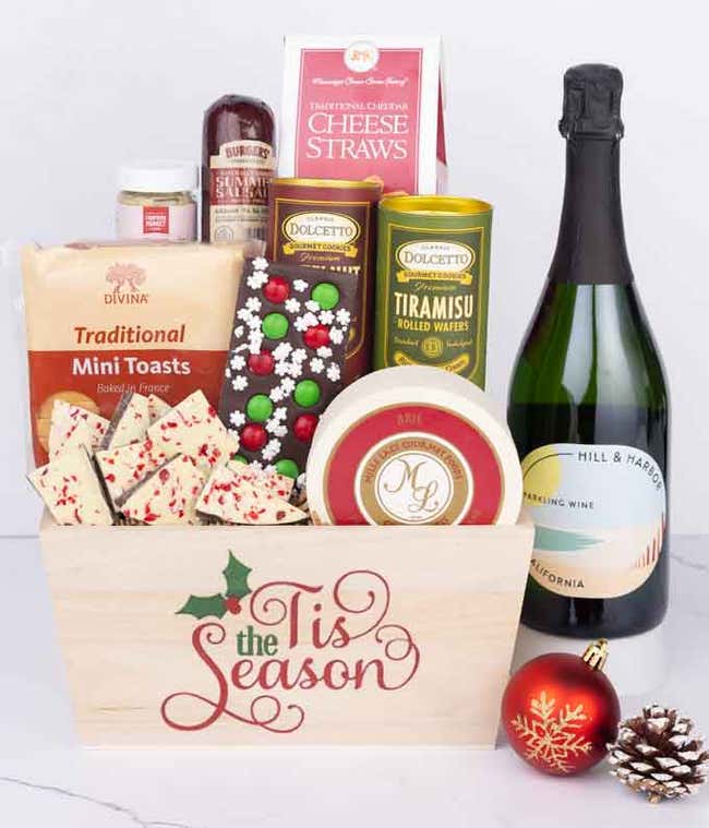 A wooden basket with the words Tis the Season, filled with sausage, cheese, mustard, cookies, wafers, and holiday bark, paired with a bottle of Hill &amp; Harbor Sparkling Wine, displayed with a red ornament and pinecone.