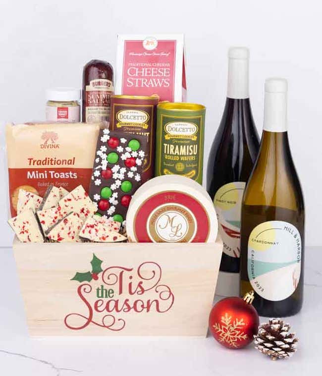 A wooden basket with the words Tis the Season, filled with sausage, cheese, mustard, cookies, toasts, wafers, and festive bark, paired with bottles of Pinot Noir and Chardonnay, with a holiday ornament and pinecone.