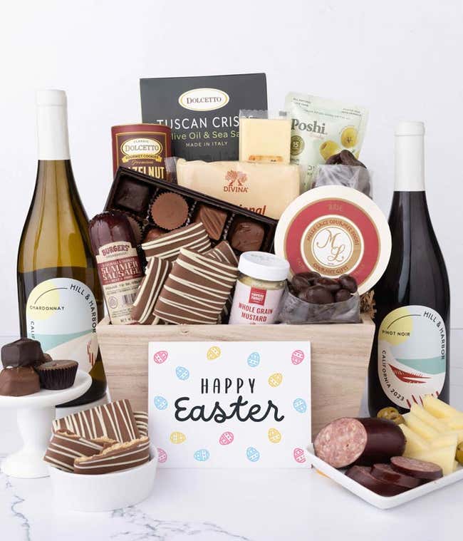 A wooden crate filled with gourmet snacks including sausage, assorted cheeses, crackers, olives, mustard, chocolate almonds, pretzels, graham crackers, assorted chocolates, and wafers, with 2 bottles of wine and an Easter card.