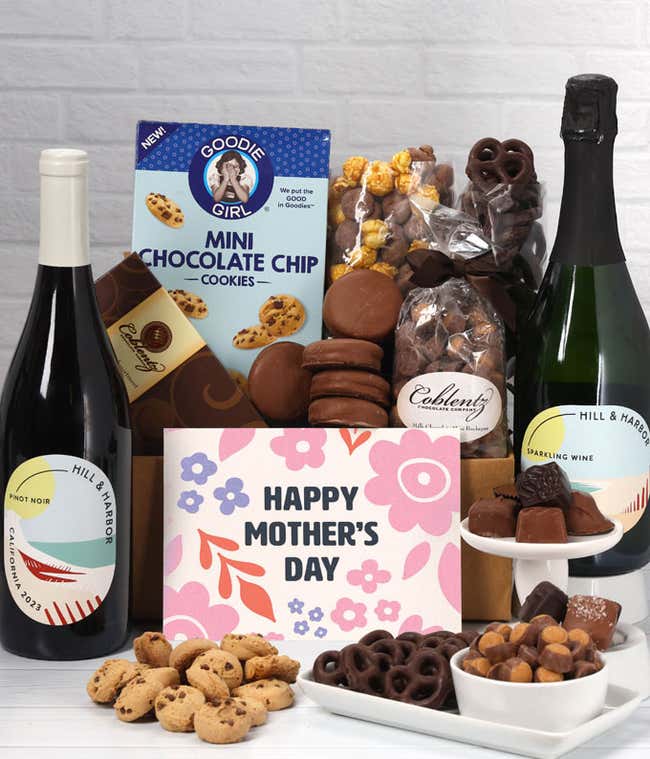 Two bottles of wine - red &amp; sparkling, sit beside chocolate-covered pretzels, mini chocolate chip cookies, caramel popcorn, assorted chocolates, mini buckeyes, chocolate covered Oreos, and a Happy Mother's Day card.
