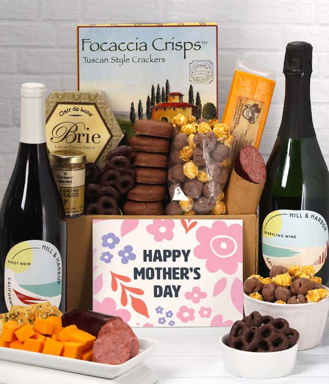 A spread featuring two bottles of wine, 2 cheeses, summer sausage, crackers, chocolate pretzels, caramel corn, and chocolate covered Oreos with a 'Happy Mother's Day' card.