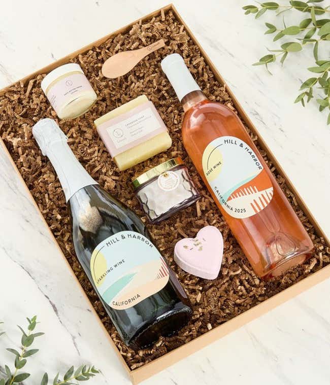 Two bottles of Hill &amp; Harbor wines displayed with spa essentials including artisan soap, bath salts with wooden scoop, and a scented candle arranged on a white surface, creating an elegant wine and self-care gift set.