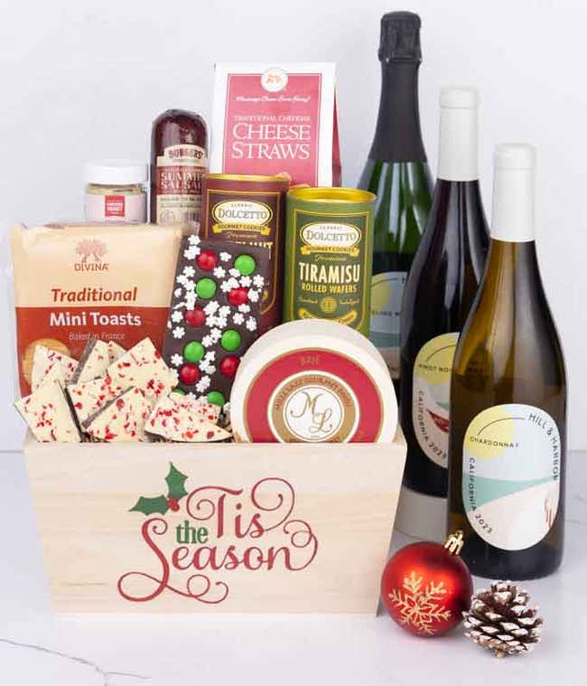 A wooden basket with the words Tis the Season, filled with sausage, cheese, mustard, cookies, wafers, and festive bark, paired with Pinot Noir, Chardonnay, and Sparkling Wine, styled with a red ornament and pinecone.